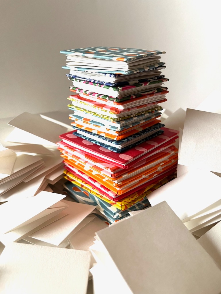 A stack of completed little folded paper books surrounded by work-in-progress of single folded squares of paper.