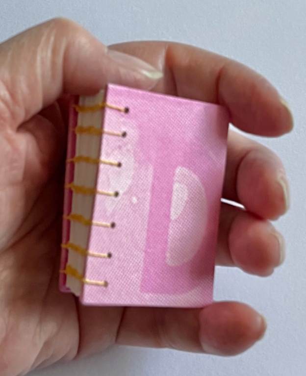 Small book with pink cover, Coptic stitch binding in yellow waxed linen thread, held in a woman's hand.